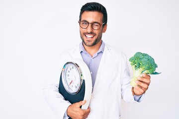 Young hispanic man as nutritionist doctor holding weighing machine and broccoli winking looking at...