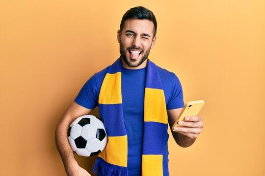 Young Hispanic Man Football Hooligan Holding Soccer Ball Using Smartphone Sticking Tongue Out Happy With Funny Expression.