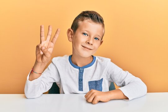 Adorable Caucasian Kid Wearing Casual Clothes Sitting On The Table Showing And Pointing Up With Fingers Number Three While Smiling Confident And Happy.