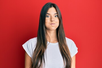 Young brunette woman wearing casual white tshirt over red background puffing cheeks with funny face. mouth inflated with air, crazy expression.
