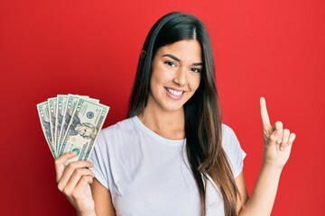 Young brunette woman holding 20 dollars banknote smiling with an idea or question pointing finger with happy face, number one