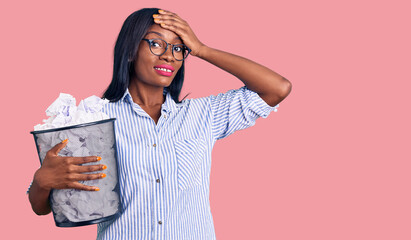 Young african american woman holding paper bin full of crumpled papers stressed and frustrated with hand on head, surprised and angry face