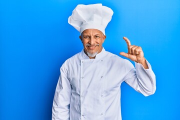 Middle age grey-haired man wearing professional cook uniform and hat smiling and confident gesturing with hand doing small size sign with fingers looking and the camera. measure concept.