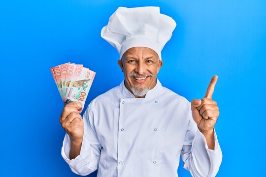 Middle age grey-haired man wearing professional cook uniform holding new zealand dollars banknotes smiling happy pointing with hand and finger to the side