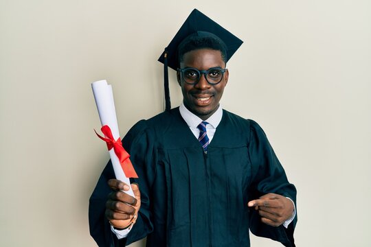 Handsome black man wearing graduation cap and ceremony robe holding diploma smiling happy pointing with hand and finger