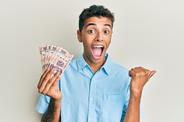 Young handsome african american man holding 500 mexican pesos banknotes pointing thumb up to the side smiling happy with open mouth