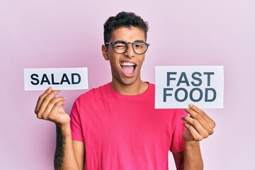 Young handsome african american man holding salad and fast food message paper winking looking at...