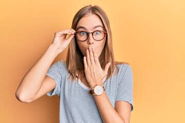 Beautiful blonde woman wearing casual clothes and holding glasses covering mouth with hand, shocked and afraid for mistake. surprised expression