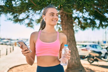 Young blonde sportswoman using smartphone and drinking bottle of water at the city.