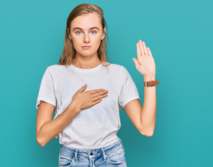 Beautiful young blonde woman wearing casual white t shirt swearing with hand on chest and open...