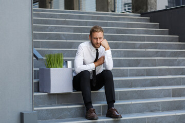 An unhappy young manager in a white shirt is fired from his job. A sad worker sits on the steps of a business center after layoffs. Crisis and unemployment.