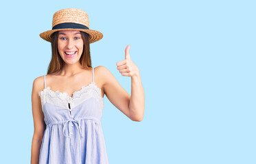 Beautiful brunette young woman wearing summer hat smiling happy and positive, thumb up doing excellent and approval sign