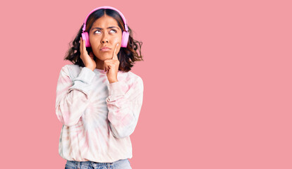 Young beautiful mixed race woman listening to music using headphones serious face thinking about...