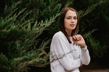 Naklejka premium Portrait of a brunette girl in a juniper. Beautiful young woman posing for a photographer in the park leaning against the branches of plants. The girl is wearing a white sweater. Free space for text