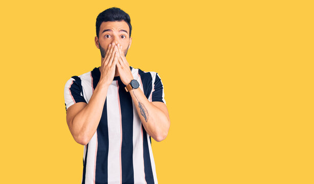 Young handsome hispanic man wearing striped tshirt shocked covering mouth with hands for mistake. secret concept.