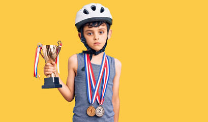 Little cute boy kid wearing bike helmet and winner medals holding winner trophy thinking attitude and sober expression looking self confident