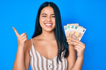 Young beautiful asian girl holding euro banknotes smiling happy pointing with hand and finger to the side