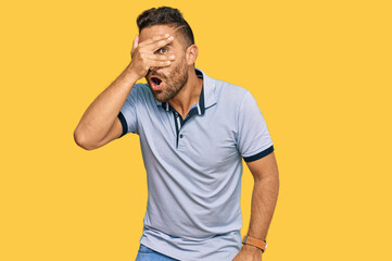 Handsome man with beard wearing casual clothes peeking in shock covering face and eyes with hand, looking through fingers with embarrassed expression.