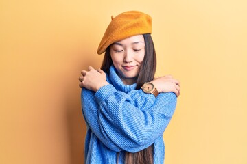 Young beautiful chinese woman wearing turtleneck sweater and french beret hugging oneself happy and positive, smiling confident. self love and self care