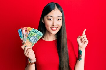 Young chinese woman holding australian dollars smiling happy pointing with hand and finger to the side