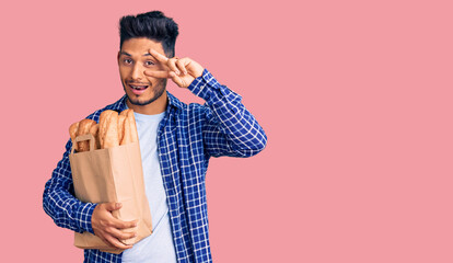 Handsome latin american young man holding paper bag with bread doing peace symbol with fingers over face, smiling cheerful showing victory