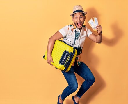 Young handsome latin man on vacation wearing summer clothes smiling happy. Jumping with smile on face holding cabin bag and airplane boarding pass over isolated background