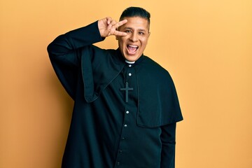 Young latin priest man standing over yellow background doing peace symbol with fingers over face, smiling cheerful showing victory