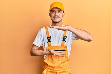 Hispanic young man wearing handyman uniform gesturing with hands showing big and large size sign, measure symbol. smiling looking at the camera. measuring concept.