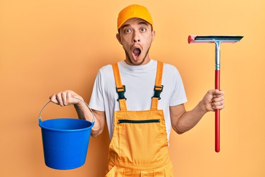 Hispanic young man wearing glass cleaner uniform and squeegee afraid and shocked with surprise and amazed expression, fear and excited face.