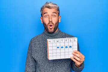 Young handsome blond man with beard holding weather calendar showing rainy week scared and amazed with open mouth for surprise, disbelief face