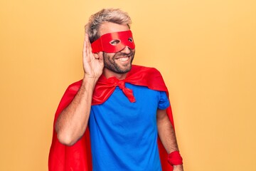 Handsome blond man wearing super hero costume with mask and cape over yellow background smiling with hand over ear listening and hearing to rumor or gossip. Deafness concept. © Krakenimages.com