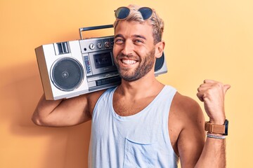 Young handsome blond man with beard wearing sunglasses listening to music using boombox pointing thumb up to the side smiling happy with open mouth © Krakenimages.com