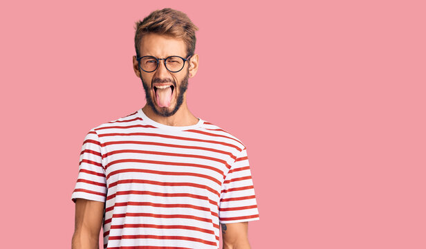 Handsome blond man with beard wearing casual clothes and glasses sticking tongue out happy with funny expression. emotion concept.