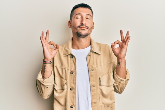 Handsome man with tattoos wearing casual brown denim jacket relax and smiling with eyes closed doing meditation gesture with fingers. yoga concept.