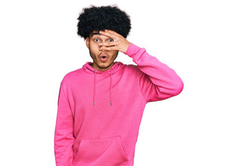 Fototapeta na wymiar Young african american man with afro hair wearing casual pink sweatshirt peeking in shock covering face and eyes with hand, looking through fingers with embarrassed expression.