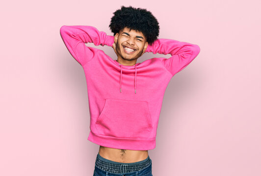 Young african american man with afro hair wearing casual pink sweatshirt relaxing and stretching, arms and hands behind head and neck smiling happy