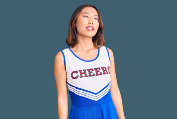 Young beautiful chinese girl wearing cheerleader uniform looking away to side with smile on face,...