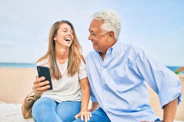 Middle age hispanic couple using smartphone sitting on the bench at the beach.