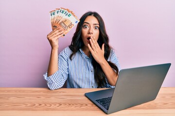 Beautiful brunette young woman working at the office holding euros covering mouth with hand,...