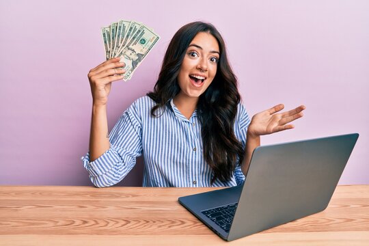 Beautiful brunette young woman working at the office holding dollars celebrating achievement with happy smile and winner expression with raised hand