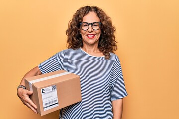 Middle age beautiful woman holding cardboard package with label over yellow background looking...