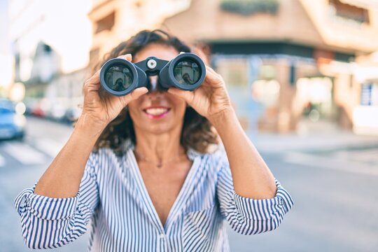 Middle age hispanic woman smiling happy looking for new opportunity using binoculars at the city.