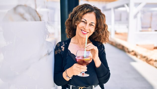 Middle age beautiful brunette woman smiling happy and confident. Leaning on the wall with smile on face drinking cup of sangria at the terrace restaurant