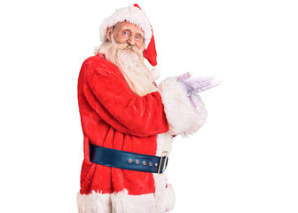 Old senior man with grey hair and long beard wearing traditional santa claus costume pointing aside with hands open palms showing copy space, presenting advertisement smiling excited happy