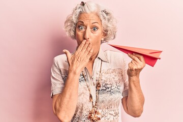 Senior grey-haired woman holding paper airplane covering mouth with hand, shocked and afraid for...
