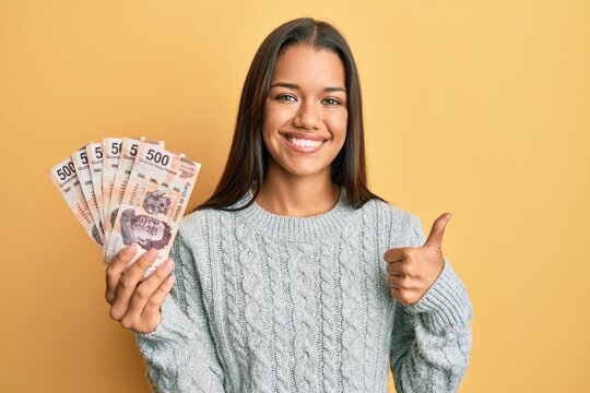 Beautiful hispanic woman holding 500 mexican pesos banknotes smiling happy and positive, thumb up doing excellent and approval sign