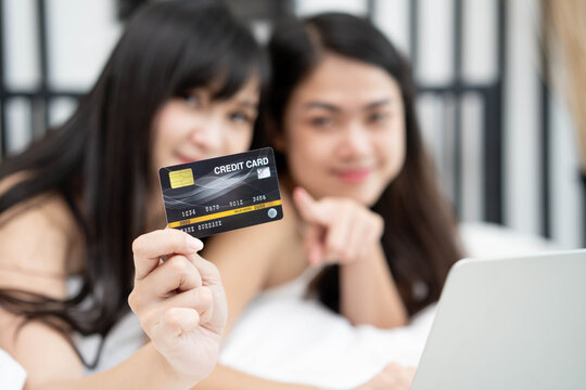 Two of happy asian young female friends surfing the internet for online shopping together on a laptop and hold credit card mockup as they laying in bed room