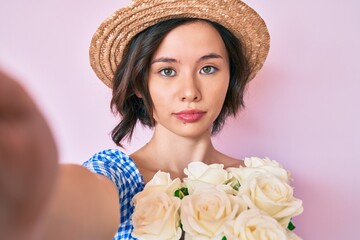 Young beautiful girl wearing summer hat taking a selfie relaxed with serious expression on face. simple and natural looking at the camera.