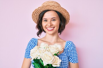 Young beautiful girl wearing summer hat holding flowers looking positive and happy standing and smiling with a confident smile showing teeth