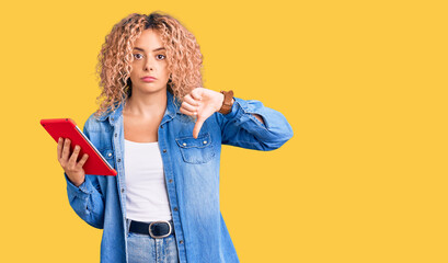 Young blonde woman with curly hair using touchpad device with angry face, negative sign showing dislike with thumbs down, rejection concept